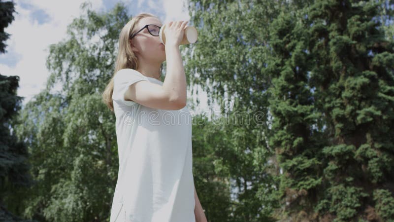 En snygg, blond flicka som dricker dryck från en bägare i parken framför fir-trädet på sommardagen. sida