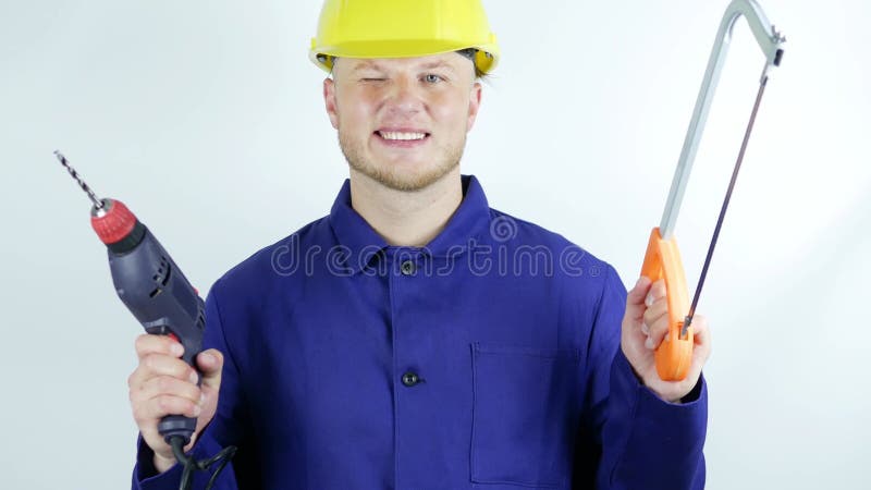 En snygg och ung byggare med en borrmaskin i händerna och en hacksåg på en vit bakgrundsleende arbetare.