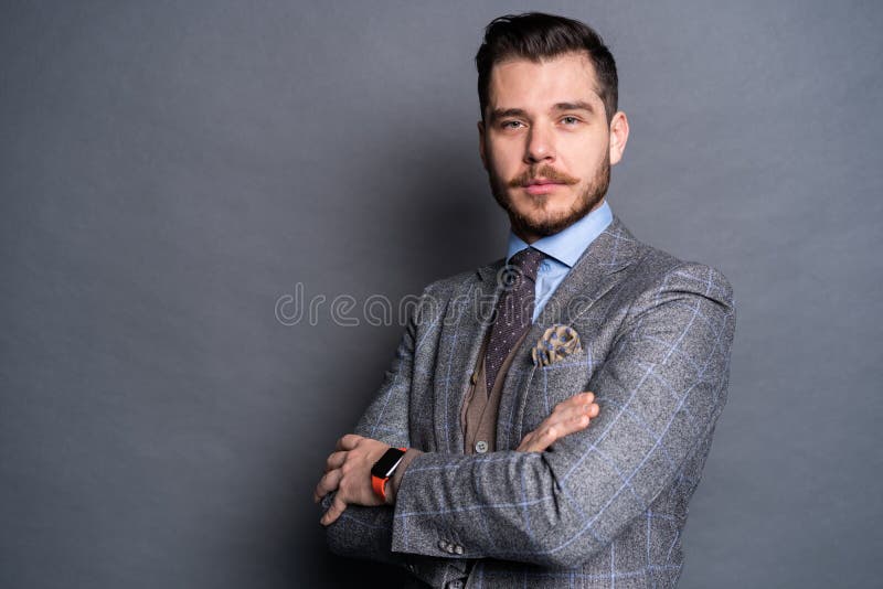 En säker elegant stilig ung man som framme står av en grå bakgrund i en studio som bär en trevlig dräkt arkivfoton En säker elegant stilig ung man som framme står av en grå bakgrund i en studio som bär en trevlig dräkt arkivfoton