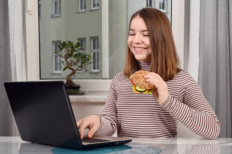 En tonåring som använder en bärbar dator och äter en vegetarisk smörgås hemma fotografering för bildbyråer
