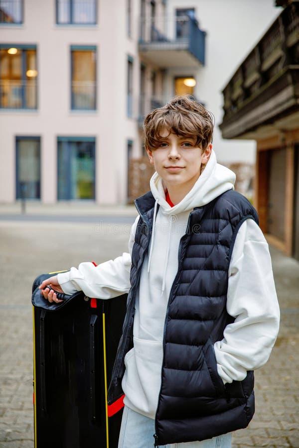 En tonårspojk med snyggt, fristående vita hodon och svart väst som står utomhus med en stor resväska redo för arkivfoto