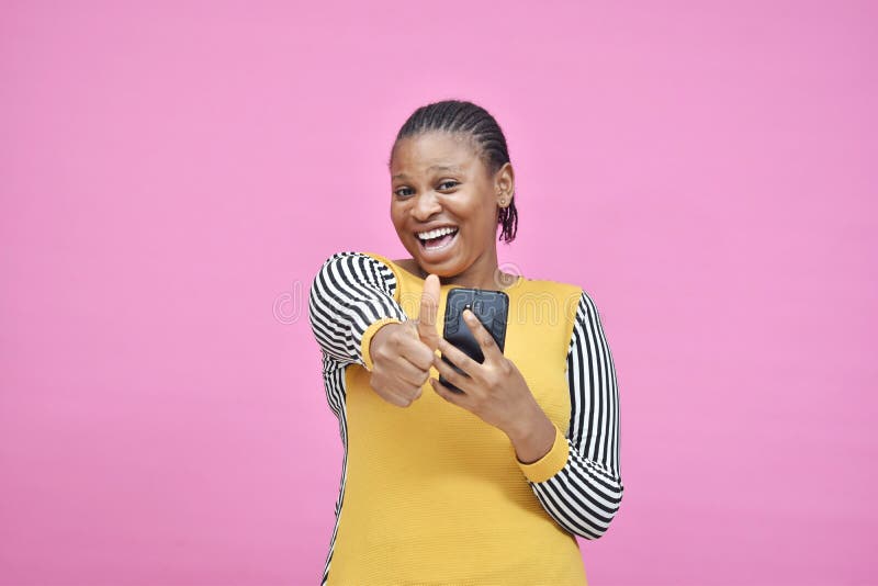 En ung afrikansk dam som håller sin mobiltelefon och ger tummen upp royaltyfri bild En ung afrikansk dam som håller sin mobiltelefon och ger tummen upp royaltyfri bild