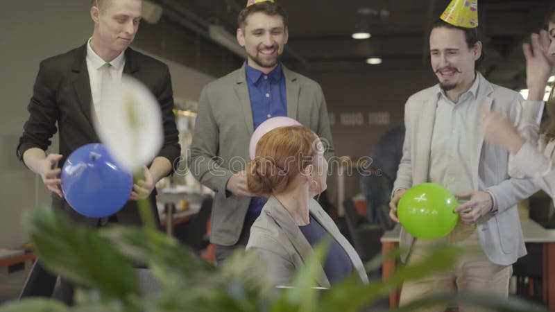 En ung, attraktiv kaukasisk kvinna som sitter vid bordet med en bärbar dator när hennes kolleger kommer med baloner på fest