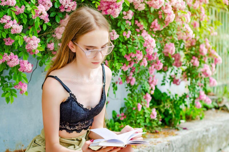 En ung blond flicka i glasögon läser en bok mot bakgrund av en blomstrande rosenbuske royaltyfria foton En ung blond flicka i glasögon läser en bok mot bakgrund av en blomstrande rosenbuske royaltyfria foton