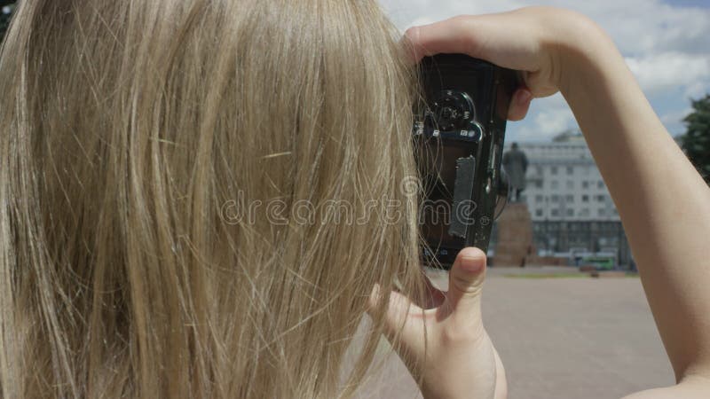 En ung blondinsk kvinna håller kamera och tar bilder på en stor parkruta och ett monument på en solig sommardag. stäng bild från