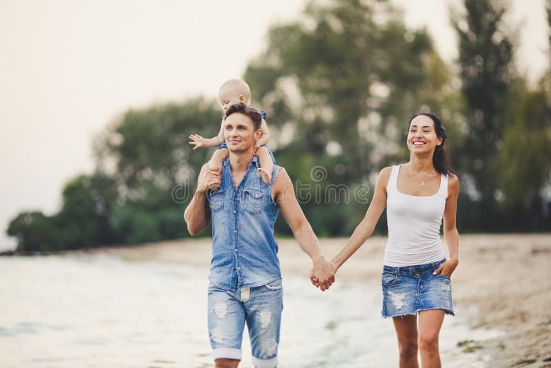 En ung familj av tre promenerar en sandig strand Behandla som ett barn lite flickan med blond som är årig från födelse, sitter på royaltyfria bilder En ung familj av tre promenerar en sandig strand Behandla som ett barn lite flickan med blond som är årig från födelse, sitter på royaltyfria bilder