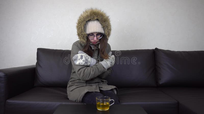 A young girl is sitting in a cold apartment on a sofa in a winter jacket. A woman with glasses tries to warm herself by hugging herself. 4k. A young girl is sitting in a cold apartment on a sofa in a winter jacket. A woman with glasses tries to warm herself by hugging herself. 4k