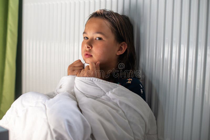 En ung flicka som sitter med en filt bredvid en radiator för att värma sig själv