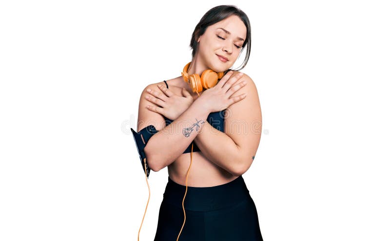 En ung historisk flicka som bär gym kläder och använder hörlurar som kramar sig själv glad och positiv och leende självsäker royaltyfri bild