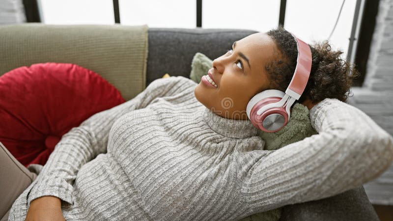 En ung kvinna leker hemma på en soffa och njuter av musik med rosa hörlurar i ett bekvämt vardagsrum arkivfoto