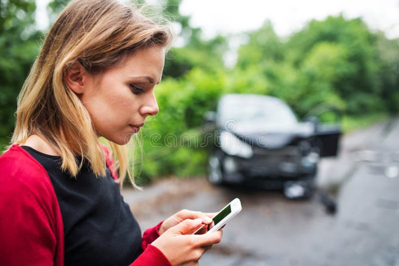 En ung kvinna med smartphonen vid den skadade bilen efter en bilolycka, textmessaging royaltyfri foto En ung kvinna med smartphonen vid den skadade bilen efter en bilolycka, textmessaging royaltyfri foto