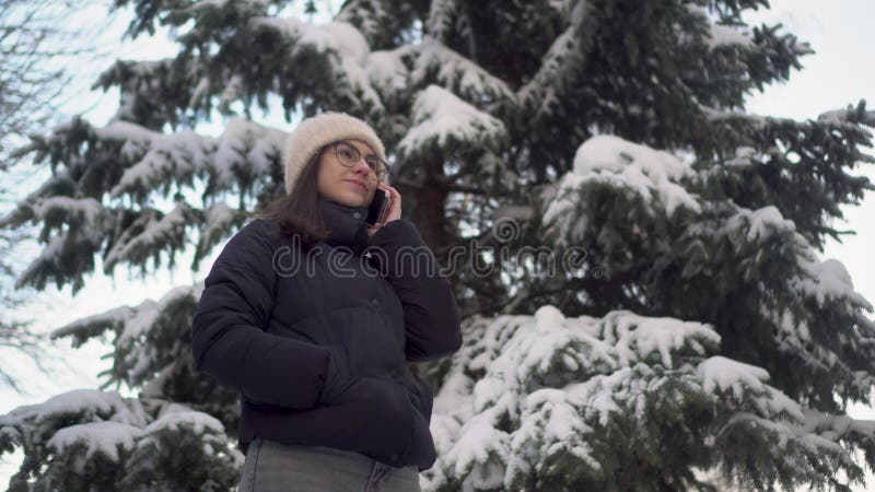 A young woman speaks on the phone near a tall spruce tree in winter. A girl with glasses communicates via cell phone against the backdrop of snowy spruce branches. 4k. A young woman speaks on the phone near a tall spruce tree in winter. A girl with glasses communicates via cell phone against the backdrop of snowy spruce branches. 4k