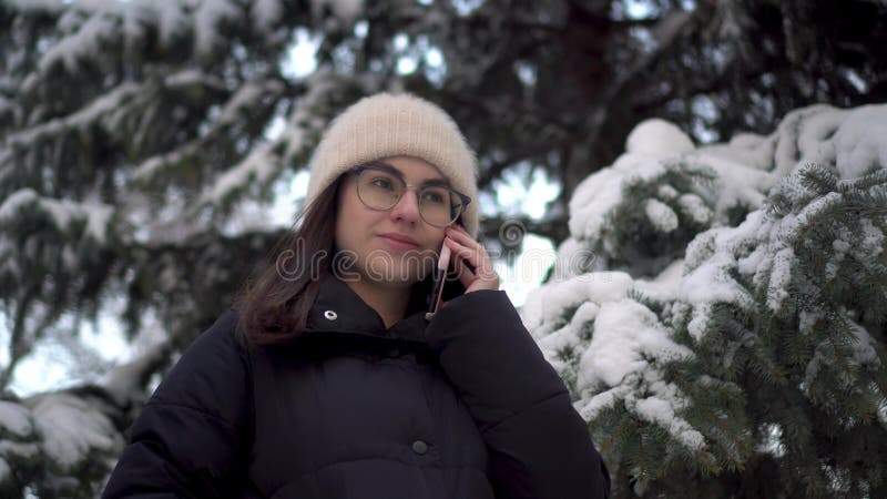 A young woman speaks on the phone near a tall spruce tree in winter. A girl with glasses communicates via cell phone against the backdrop of snowy spruce branches. 4k. A young woman speaks on the phone near a tall spruce tree in winter. A girl with glasses communicates via cell phone against the backdrop of snowy spruce branches. 4k