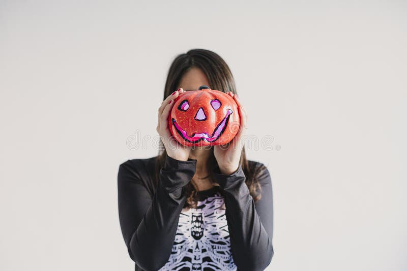 En ung kvinna som håller en pumpa som täcker hennes ansikte Med ett svart och vitt skelett Halloween-koncept Inomhus livsstil arkivbilder