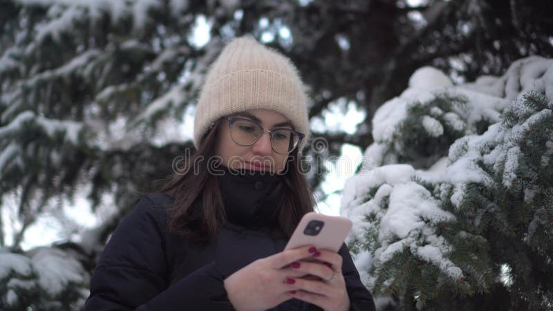 A young woman stands with a phone against a background of spruce in winter. A girl in glasses with a smartphone chats against the backdrop of snowy spruce branches. 4k. A young woman stands with a phone against a background of spruce in winter. A girl in glasses with a smartphone chats against the backdrop of snowy spruce branches. 4k