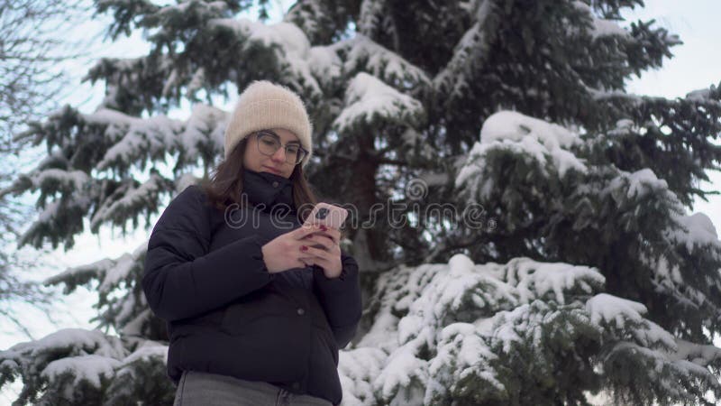 A young woman stands with a phone against a background of spruce in winter. A girl in glasses with a smartphone chats against the backdrop of snowy spruce branches. 4k. A young woman stands with a phone against a background of spruce in winter. A girl in glasses with a smartphone chats against the backdrop of snowy spruce branches. 4k