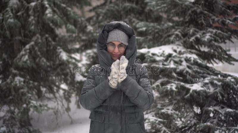 A young woman stands against a background of fir trees under heavy snowfall and rubs her hands against the cold. A girl in glasses and a down jacket with a hood stands and looks at the camera. 4k. A young woman stands against a background of fir trees under heavy snowfall and rubs her hands against the cold. A girl in glasses and a down jacket with a hood stands and looks at the camera. 4k