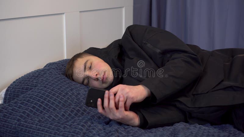 A young man in a jacket lies on the bed and uses a smartphone. The man did not pay the bills and was left without heating in the winter. 4k. A young man in a jacket lies on the bed and uses a smartphone. The man did not pay the bills and was left without heating in the winter. 4k