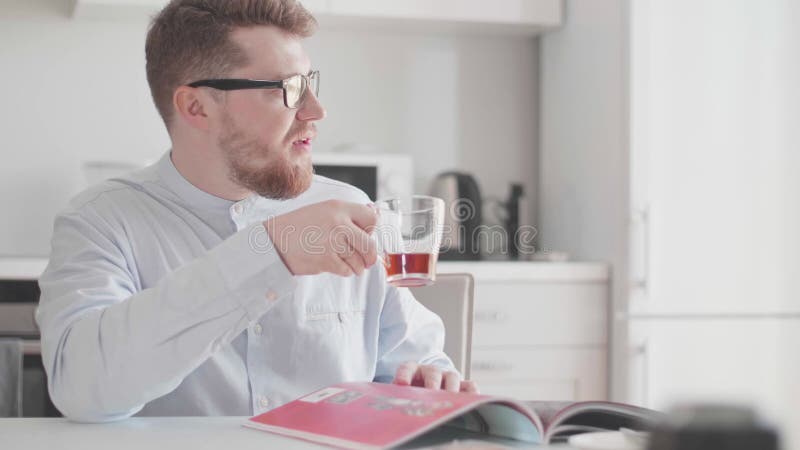 En ung, stilig man med glasögon på morgonen vid frukost läser en tidning och dricker te