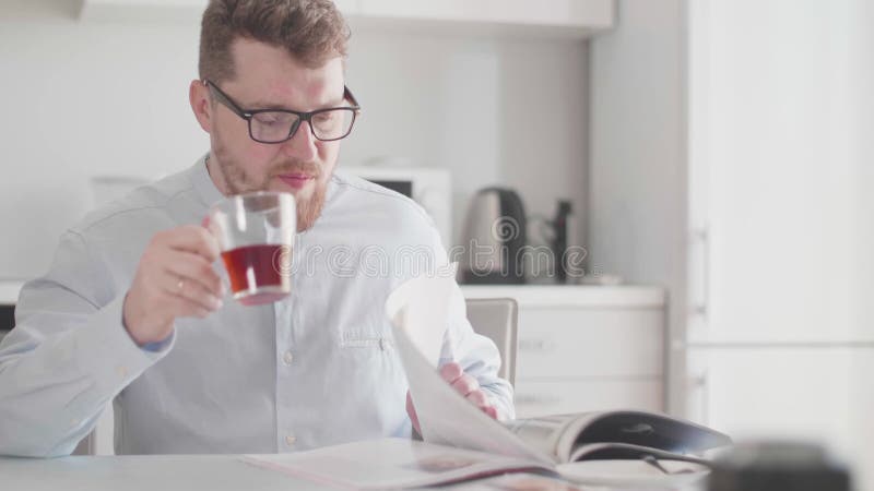 En ung, stilig man med glasögon på morgonen vid frukost läser en tidning och dricker te