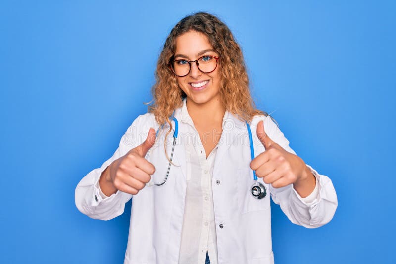 En ung, vacker blondindoktor med blå ögon som bär rock och stethoscope som godkänner att man gör en positiv gest med handtummarna. arkivbild