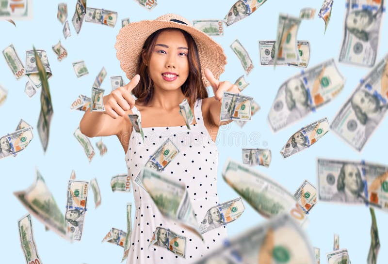 En ung vacker kinesisk flicka som bär sommarhatt och som godkänner att man gör en positiv gest med handtummarna upp och log och gl royaltyfri fotografi