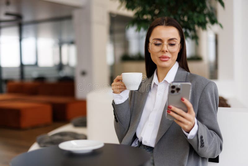 En ung, vacker kvinna håller i en kaffekopp och tittar på sin smarttelefon medan hon sitter på ett kafé arkivbild