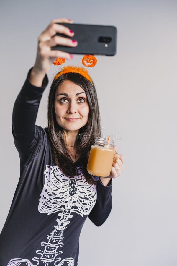 En ung vacker kvinna som dricker apelsinjuice och tar en selfie med mobiltelefon. Med ett svart och vitt skelett arkivfoto