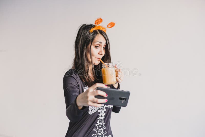 En ung vacker kvinna som dricker apelsinjuice och tar en selfie med mobiltelefon. Med ett svart och vitt skelett royaltyfri foto