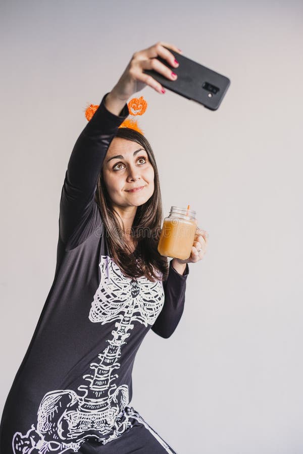 En ung vacker kvinna som dricker apelsinjuice och tar en selfie med mobiltelefon. Med ett svart och vitt skelett arkivfoto