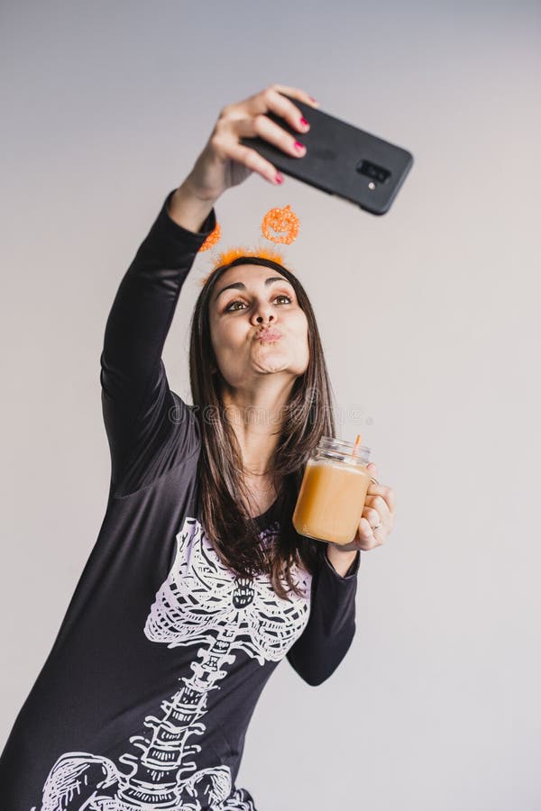 En ung vacker kvinna som dricker apelsinjuice och tar en selfie med mobiltelefon. Med ett svart och vitt skelett royaltyfri fotografi