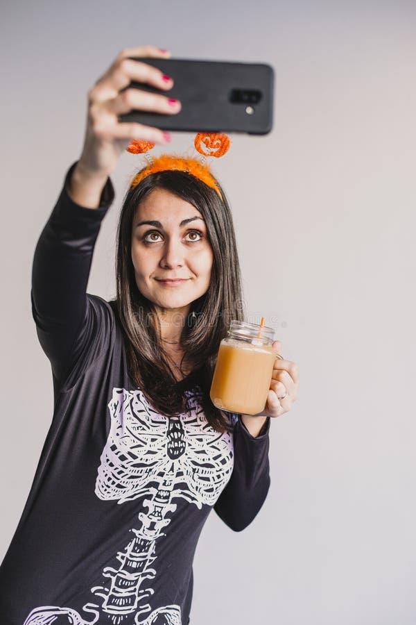 En ung vacker kvinna som dricker apelsinjuice och tar en selfie med mobiltelefon. Med ett svart och vitt skelett arkivbilder