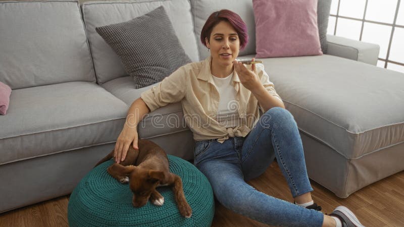 En ung vacker kvinna som sitter i ett vardagsrum i hennes hus och pratar på sin smartphone med sin hushund vilande på en dyshion royaltyfri bild