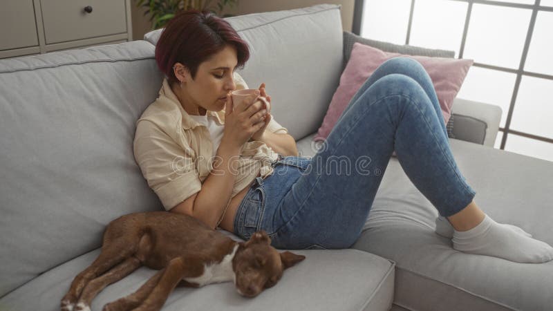 En ung vacker kvinnas relaxer på en soffa med sin hushund i ett mystiskt vardagsrum som njuter av en varm kopp te inomhus royaltyfri fotografi