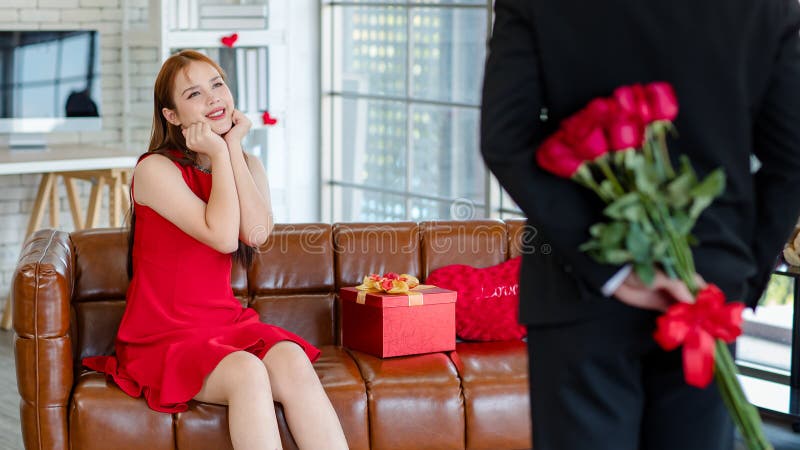 En ung, vacker kvinnlig, överraskad flickvän i den röda klänningen som leker på den galna soffan medan en okänd manlig pojkvän håller blommor bouquet bakom sig