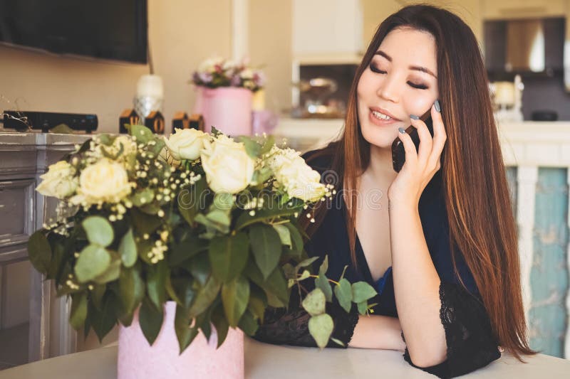 En vacker asiatisk kvinna pratar telefonplatser vid bordet i vardagsrummet... en ljus invändig beundrare av vita rosor blommor bou arkivbild