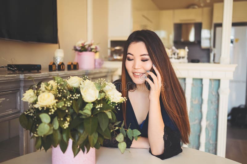 En vacker asiatisk kvinna pratar telefonplatser vid bordet i vardagsrummet... en ljus invändig beundrare av vita rosor blommor bou arkivbild