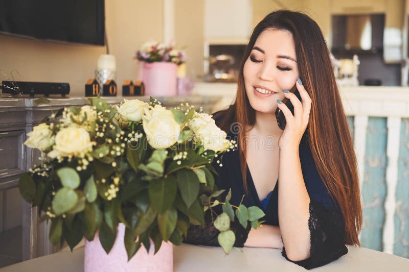 En vacker asiatisk kvinna pratar telefonplatser vid bordet i vardagsrummet... en ljus invändig beundrare av vita rosor blommor bou royaltyfria foton