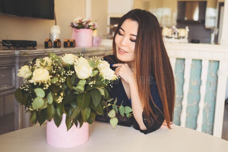 En vacker asiatisk kvinna sitter vid bordet i vardagsrummet.. -... beundrar vita rosor blommor bouquet.. leende royaltyfri foto