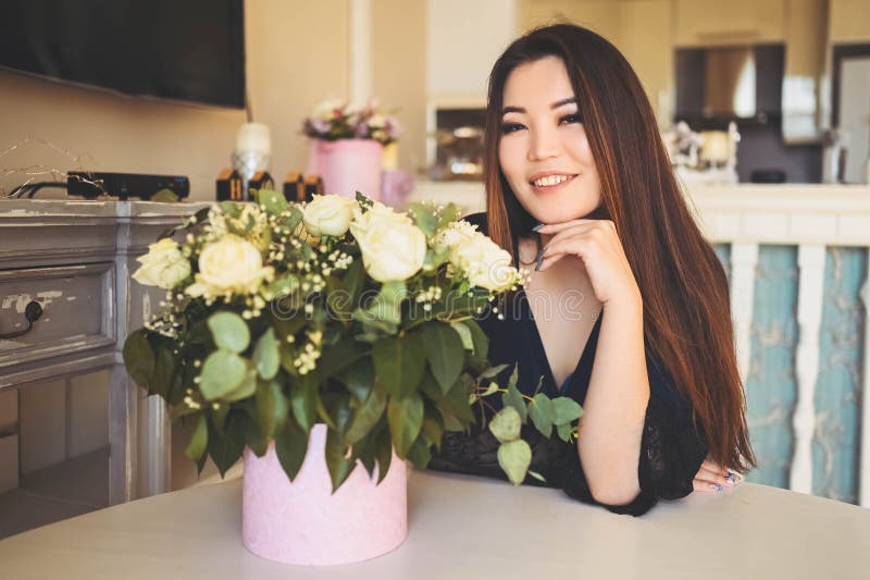 En vacker asiatisk kvinna sitter vid bordet i vardagsrummet.. -... beundrar vita rosor blommor bouquet.. leende royaltyfria bilder