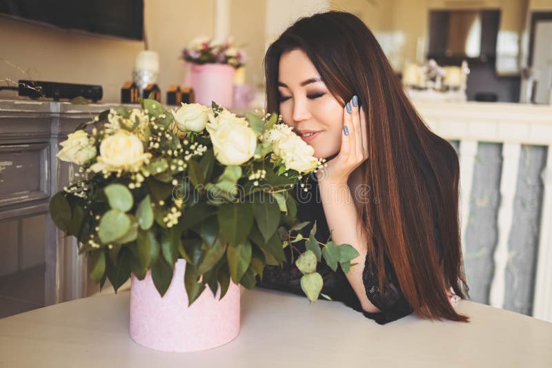 En vacker asiatisk kvinna sitter vid bordet i vardagsrummet.. -... beundrar vita rosor blommor bouquet.. leende royaltyfri foto
