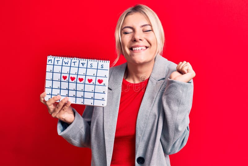 En vacker blond kvinna som kontrollerar menstruationstid i kalendern över röd bakgrund skriker stolt över segern fotografering för bildbyråer