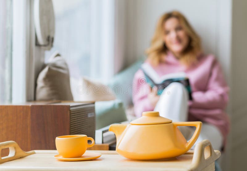 A beautiful woman in a pink hoodie sits on the windowsill, reads a book and drinks tea. Focus on teapot and cup. Slow life concept. A beautiful woman in a pink hoodie sits on the windowsill, reads a book and drinks tea. Focus on teapot and cup. Slow life concept