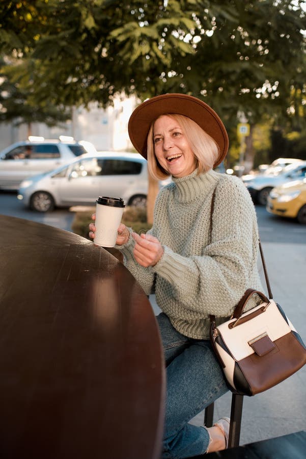 Beautiful mature woman 50-55 year old drink coffee in paper cup wear knit sweater and felt hat  sitting in cafe outdoors. Autumn season. Healthy aged blonde female. Happiness. Beautiful mature woman 50-55 year old drink coffee in paper cup wear knit sweater and felt hat  sitting in cafe outdoors. Autumn season. Healthy aged blonde female. Happiness