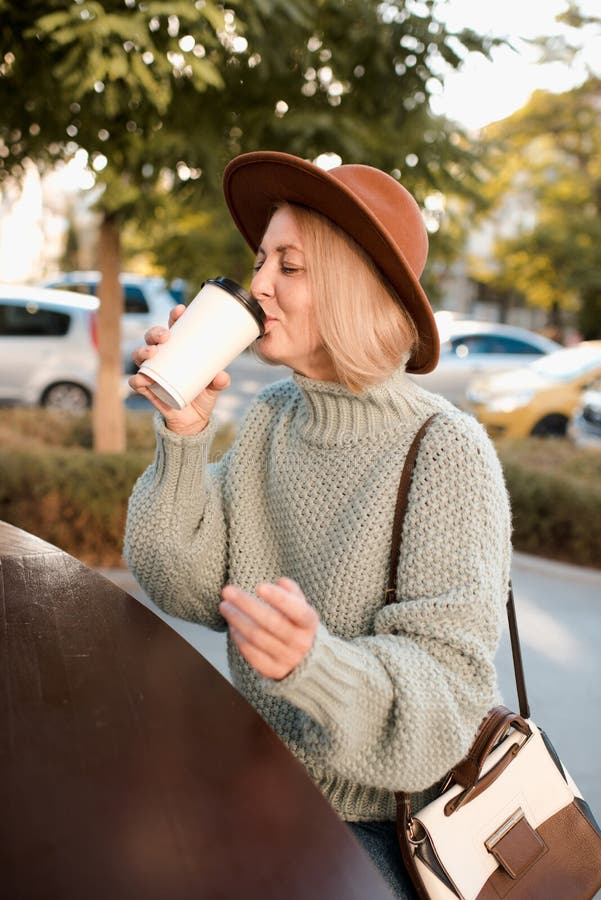 Beautiful mature woman 50-55 year old drink coffee in paper cup wear knit sweater and felt hat sitting in cafe outdoors. Autumn season. Healthy aged blonde female. Happiness. Beautiful mature woman 50-55 year old drink coffee in paper cup wear knit sweater and felt hat sitting in cafe outdoors. Autumn season. Healthy aged blonde female. Happiness