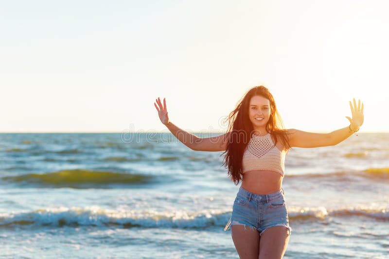 En vacker sexig flicka som höjer upp händerna på havsstranden i en tillfällig klädsel tittar på kameran vid solnedgången royaltyfri fotografi