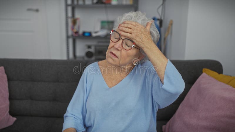 En äldre kvinna med grå hår och glasögon framstår som stressad och orolig i ett glatt vardagsrum royaltyfri foto