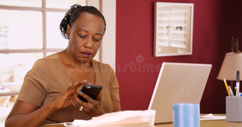 En äldre svart kvinna använder hennes telefon och bärbar dator för att göra henne skatter