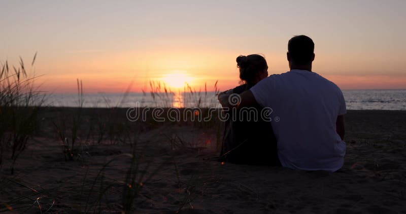 Enamorados de la pareja sentados en la arena en la playa al romántico atardecer dorado. vista posterior almacen de video