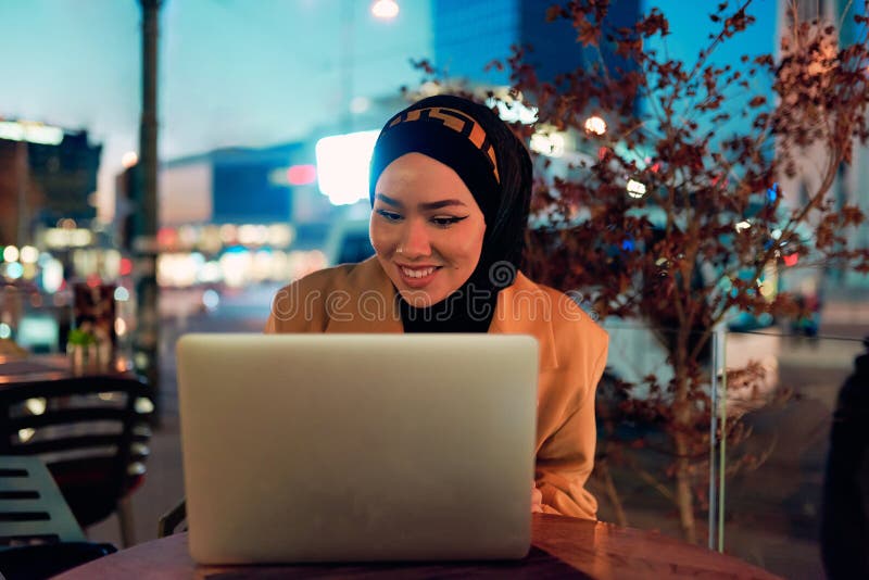 în atmosfera încântătoare a unui peisaj nocturn al orașului, o hijabclad-girl îngropată în laptop-ul ei crează o fascinantă stock photography